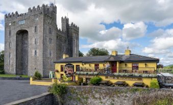 a yellow building with a red door and a tall tower in the background , surrounded by trees and grass at Bunratty Castle Hotel