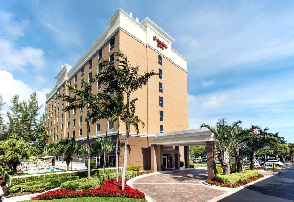 a hampton inn hotel with a large sign , surrounded by palm trees and flowers , under a clear blue sky at Hampton Inn Hallandale Beach-Aventura