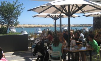 a group of people sitting under umbrellas at an outdoor dining area near a body of water at The Marina Hotel - Mindarie