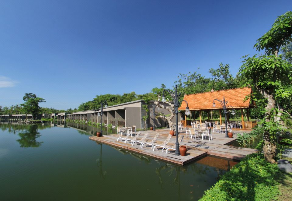 a serene lake with a wooden dock extending into the water , surrounded by trees and buildings at The Westlake Hotel & Resort Yogyakarta