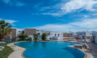 a large , circular swimming pool surrounded by white buildings and blue skies , with trees and umbrellas visible in the area at Naxos Palace Hotel