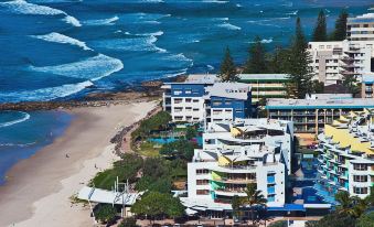 a beach scene with a hotel building situated on the edge of the ocean , surrounded by trees and sand at Rolling Surf Resort Sunshine Coast