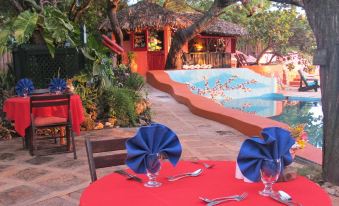 a restaurant with red tables and blue napkins , surrounded by lush greenery and a pool in the background at Hope House