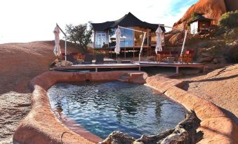 a large pool is surrounded by a red rock wall and has a black tent in the background at Spitzkoppen Lodge