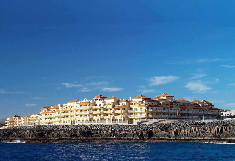 a large building is situated on a rocky shoreline , with a blue sky and ocean in the background at Elba Castillo San Jorge & Antigua Suite Hotel