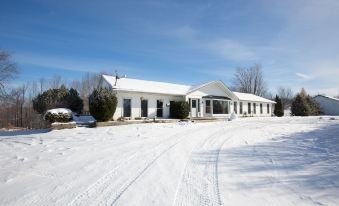 a white building with a sloping roof , surrounded by snow - covered trees and cars parked on the street at White House Farm