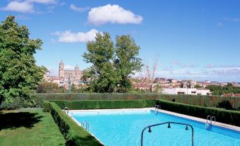 a large outdoor swimming pool surrounded by grass , trees , and a city skyline in the background at Parador de Salamanca