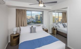 a modern bedroom with a large window , white walls , and blue and white bedding , as well as a wooden desk and a fan at Direct Collective - Pavilion and Governor on Brookes