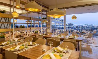 a restaurant with wooden tables and chairs , yellow pendant lights , and a view of the ocean at Zafiro Palace Alcudia