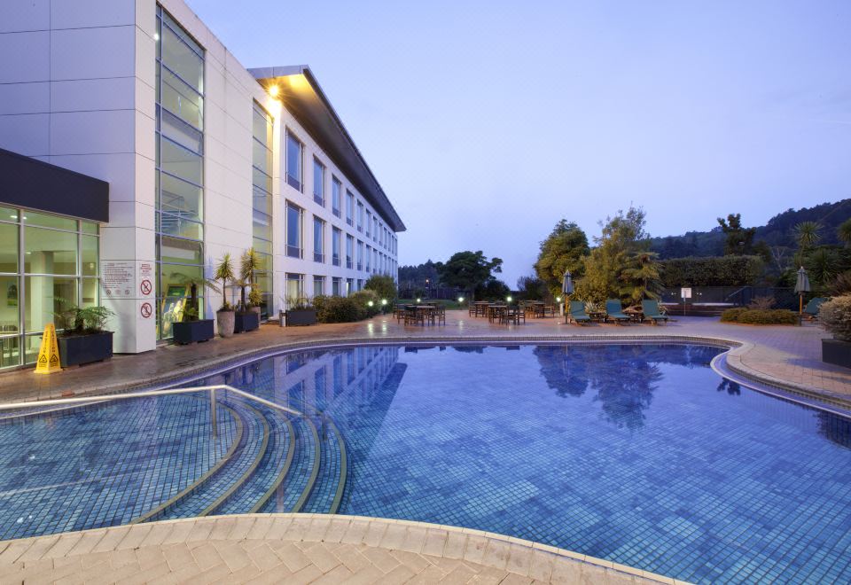 an outdoor swimming pool surrounded by a hotel building , with the sun setting in the background at Rydges Rotorua, an EVT Hotel