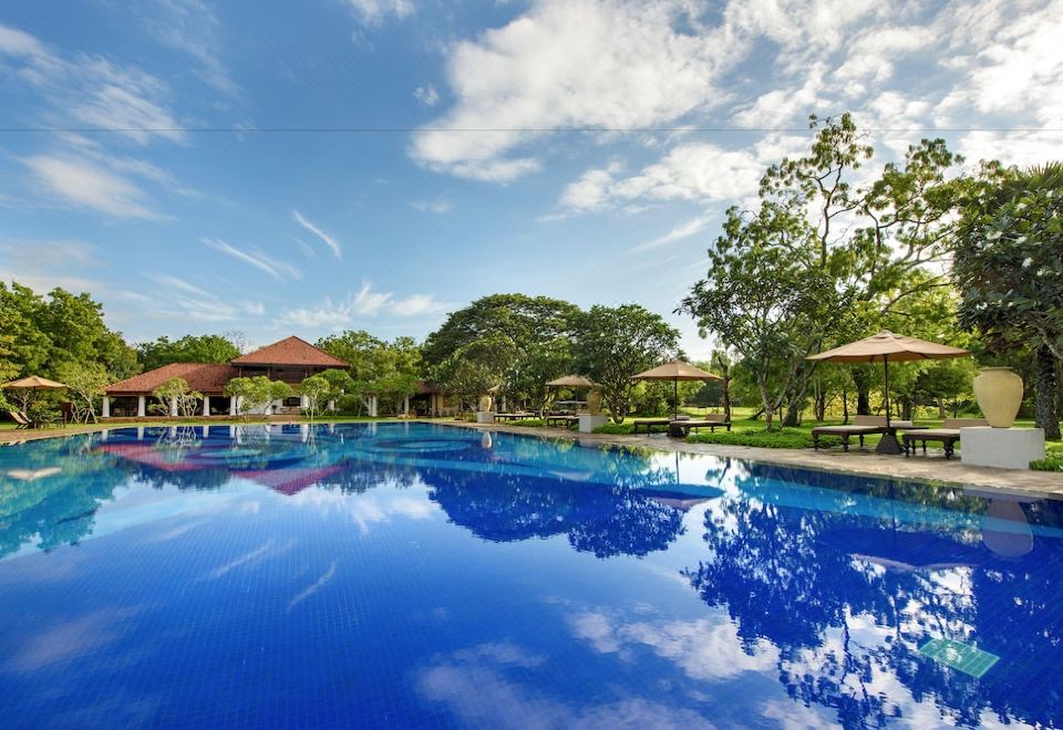 a large blue swimming pool surrounded by green trees and grass , with umbrellas and benches placed around it at Uga Ulagalla