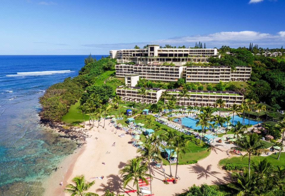 aerial view of a resort with a beach , palm trees , and a large building on the cliff at 1 Hotel Hanalei Bay