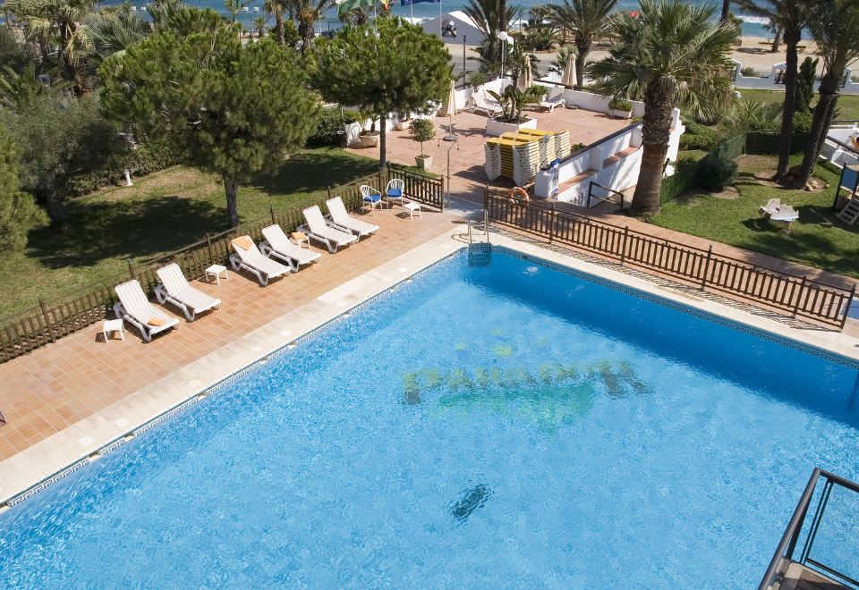 a large outdoor swimming pool surrounded by lounge chairs and palm trees , with the ocean visible in the background at Parador de Mojacar