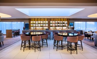 a modern bar with wooden chairs and tables , surrounded by a glass window and shelves filled with various bottles at Leonardo Hotel and Conference Venue Hinckley Island