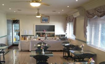 a dining area with tables , chairs , and a tv . a kitchen in the background , and a flower arrangement on the at Crest Motor Inn