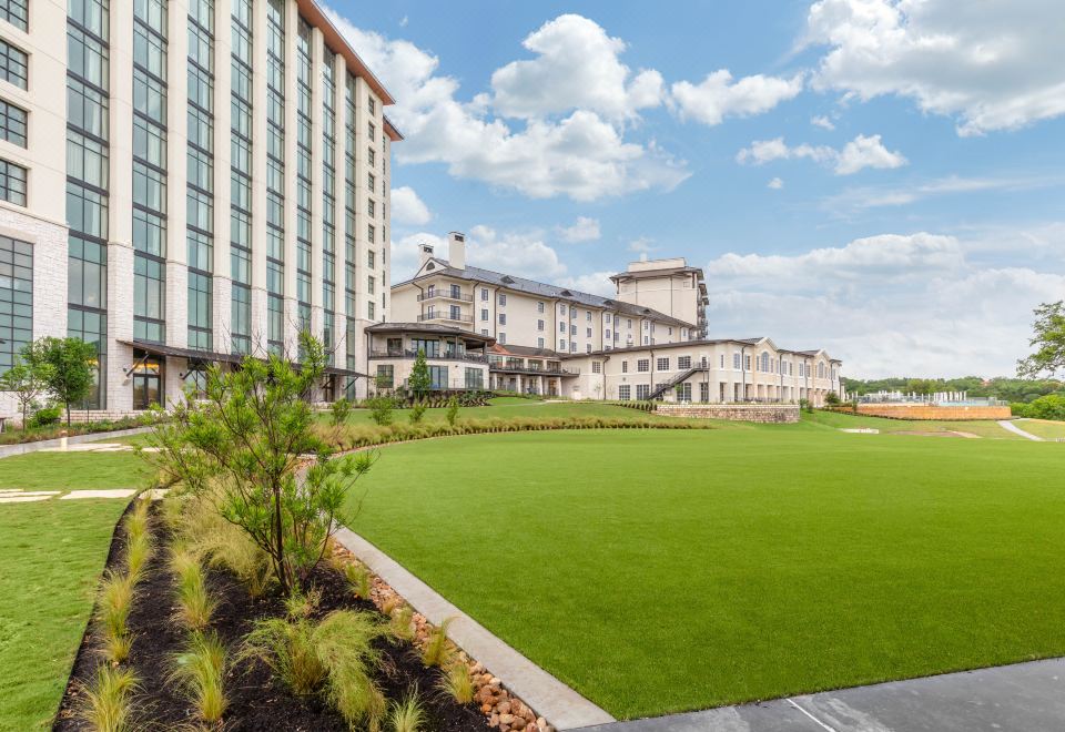 a large hotel with a grassy lawn and tall buildings in the background , under a blue sky with clouds at Omni Barton Creek Resort and Spa Austin