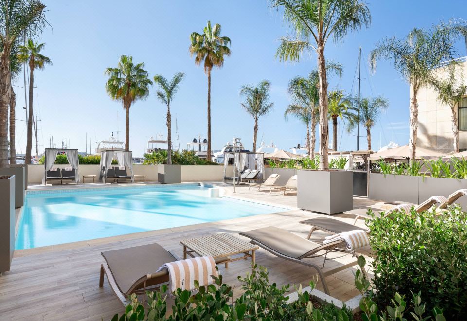a large outdoor swimming pool surrounded by palm trees , with lounge chairs and umbrellas placed around the pool area at Riviera Marriott Hotel la Porte de Monaco
