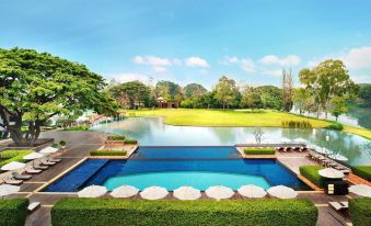 a large outdoor swimming pool surrounded by lush greenery , with a lake in the background at Le Meridien Chiang Rai Resort, Thailand