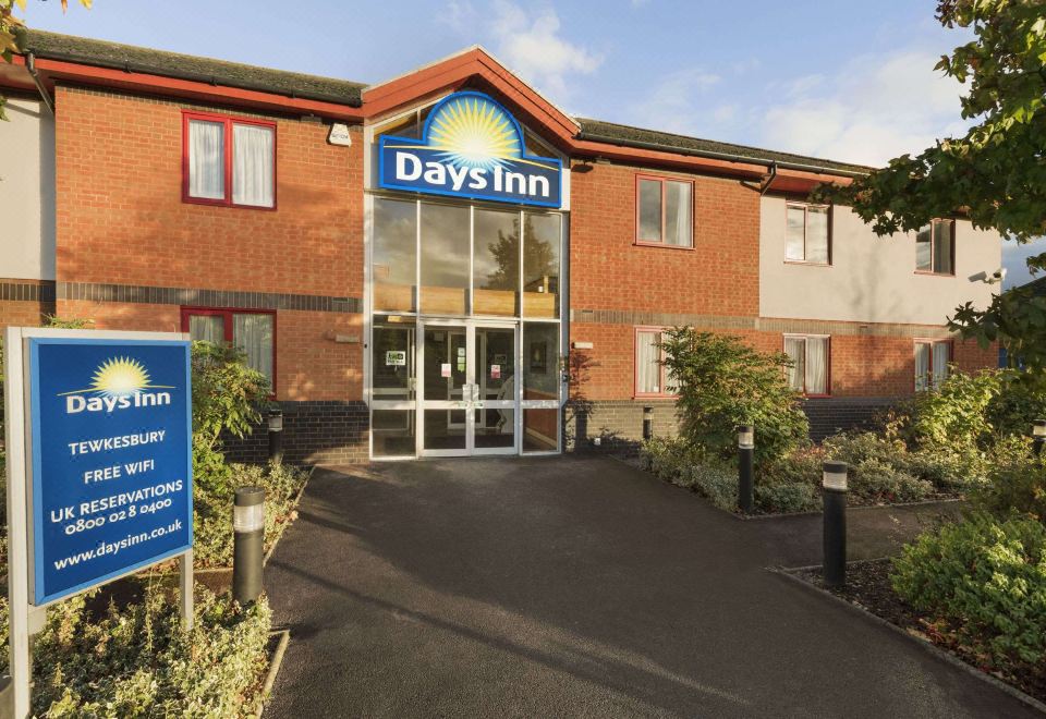 a days inn hotel with its sign , surrounded by trees and bushes , under a clear blue sky at Days Inn by Wyndham Tewkesbury Strensham