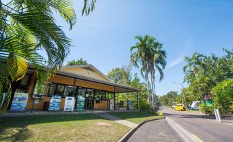 a tropical setting with a yellow bus parked in front of a building , surrounded by palm trees and a blue sky at Hidden Valley Holiday Park