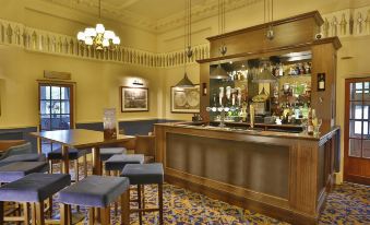 a bar with a wooden counter and several stools , featuring a chandelier hanging from the ceiling at Reigate Manor Hotel