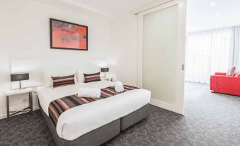 a large bed with white linens and a red and black striped blanket is in a hotel room at City Edge Box Hill Apartment Hotel