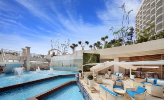 a luxurious resort with a large pool surrounded by palm trees , chairs , and umbrellas for guests to relax and enjoy their time at Loisir Hotel Naha