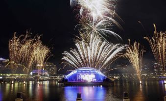 a nighttime scene with a large group of people gathered on the bank of a body of water , watching a fireworks display at Novotel Sydney Darling Harbour