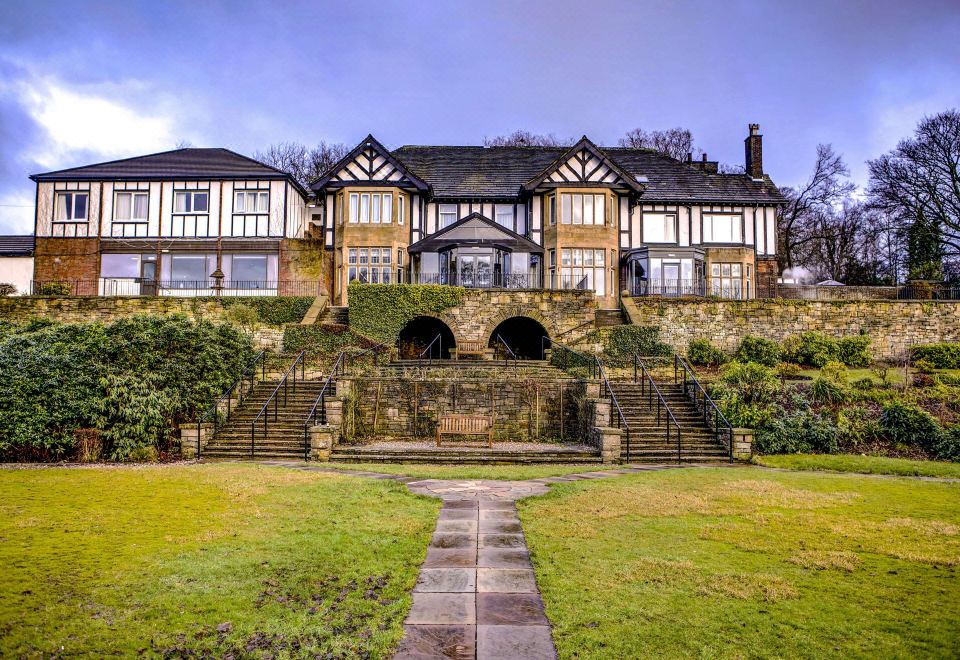 a large , two - story house with a stone facade and wooden balconies is surrounded by a well - maintained garden at Burnley West Higher Trapp Hotel