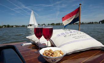 a wooden boat is docked with a sailboat in the background , and two wine glasses and a bowl of snacks on the table at Fletcher Hotel Restaurant Loosdrecht-Amsterdam