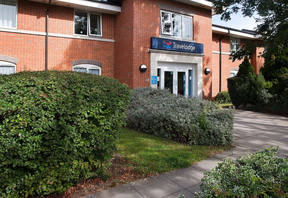 a brick building with a blue sign on the front , surrounded by bushes and trees at Travelodge Stonehouse