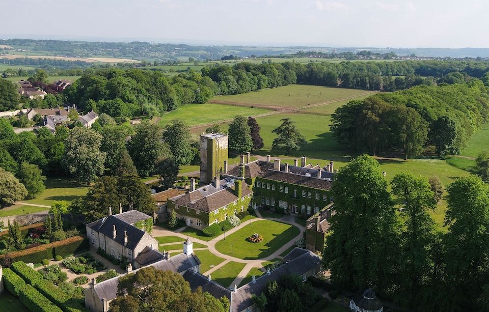 an aerial view of a large , green building surrounded by trees and grass , with a road leading to it at Lucknam Park Hotel