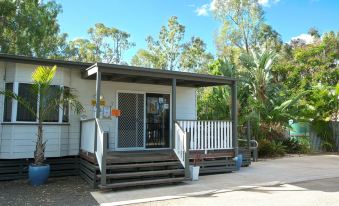 a small , two - story house with a wooden deck and white railings , surrounded by trees on a sunny day at Discovery Parks – Biloela