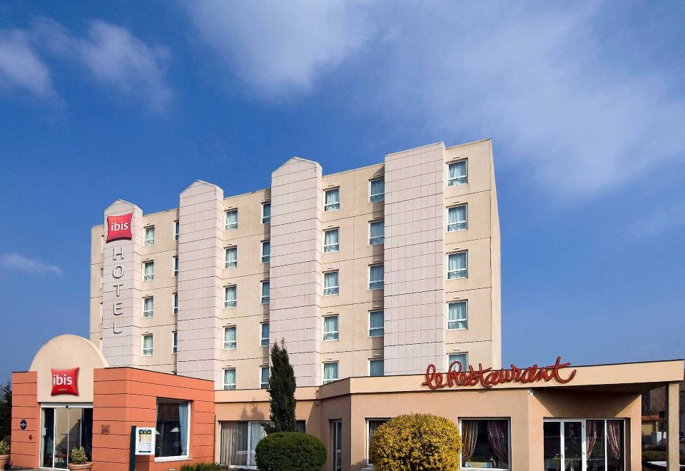 a hotel with a red sign and beige exterior , under a clear blue sky with white clouds at Ibis Clermont-Ferrand Sud Carrefour Herbet