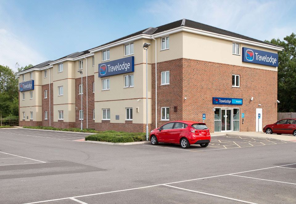 a red car is parked in a parking lot outside of a two - story building with blue signage at Travelodge Wincanton