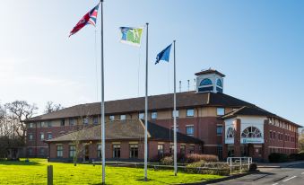 "a large brick building with multiple flags flying outside , including one that says "" cool white "", and three smaller flags" at Holiday Inn Express Warwick - Stratford-Upon-Avon