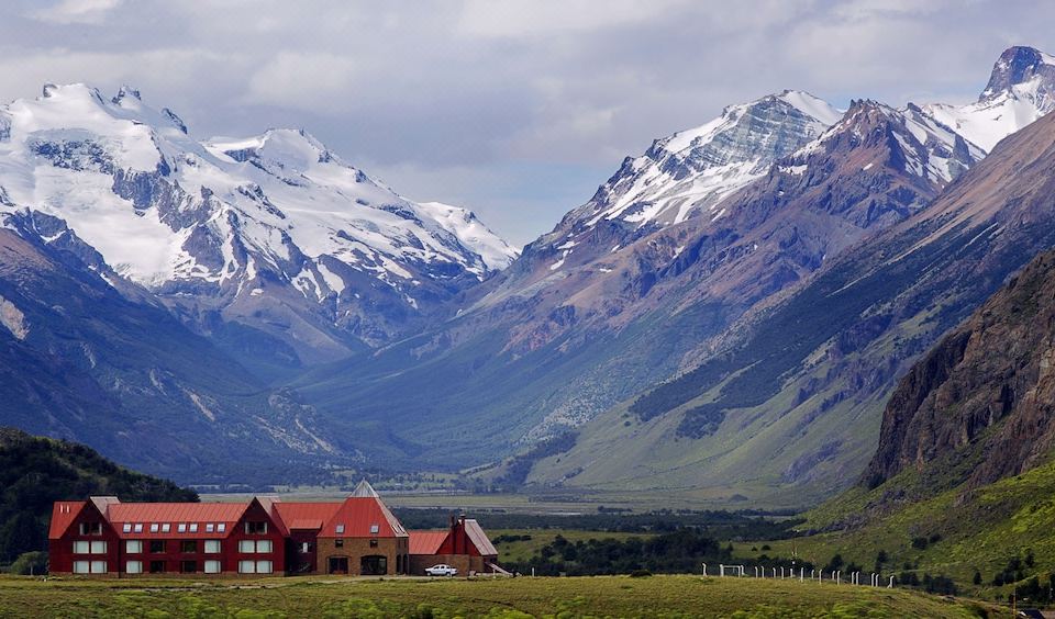 a red house is nestled in a valley with snow - capped mountains in the background , and green fields in the foreground at Los Cerros del Chalten Boutique Hotel