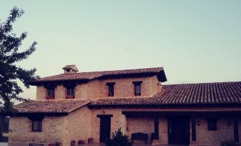 a large , two - story house with a red tile roof and a stone wall is surrounded by potted plants at La Contrada