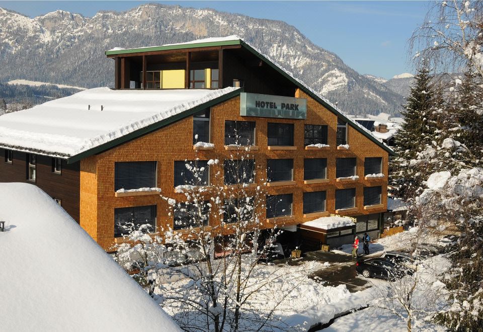 a large wooden building with a green roof is nestled in the snow , with mountains visible in the background at Hotel Park