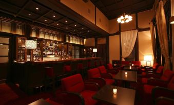 a dimly lit bar with red chairs and tables , a chandelier hanging from the ceiling , and a long counter at Nara Hotel