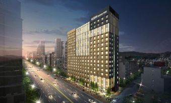 a modern hotel building with a large glass facade , surrounded by trees and a busy city street at dusk at Lotte City Hotel Ulsan