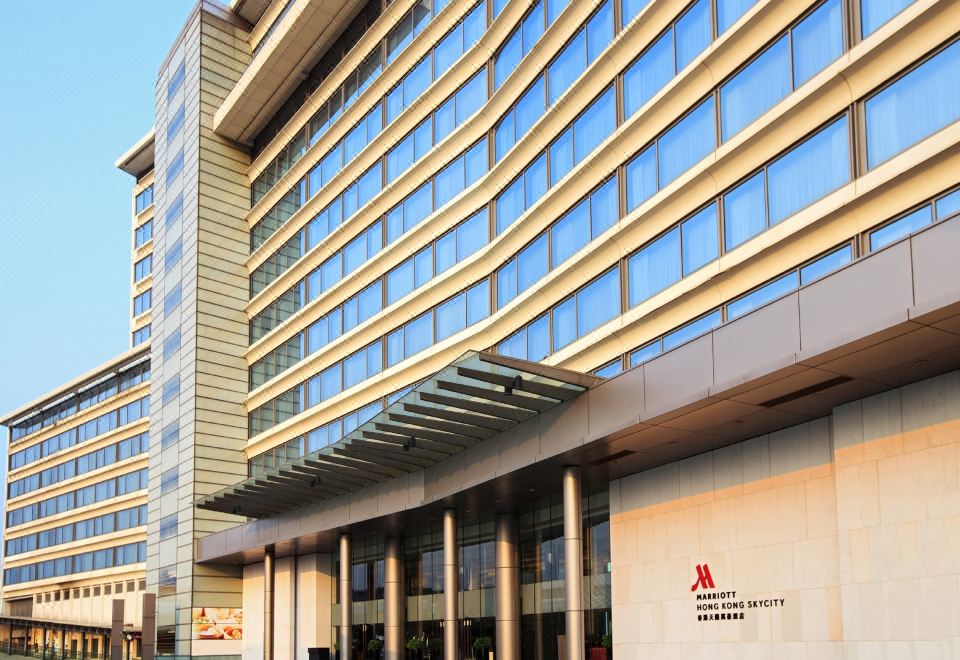 A large building's front entrance is depicted, with an outside view and other buildings in the background at Hong Kong SkyCity Marriott Hotel