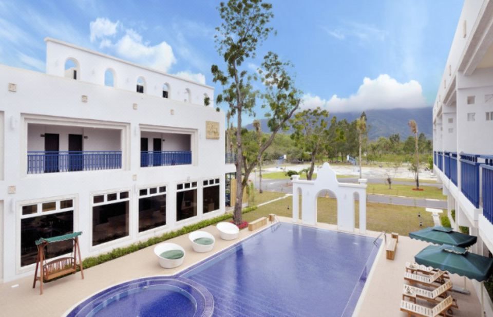 a white building with a large pool in front of it , surrounded by palm trees and mountains in the background at J Resort