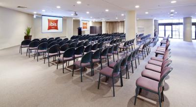 a large conference room with rows of chairs arranged in a semicircle , and a projector screen on the wall at Ibis Melbourne Hotel and Apartments