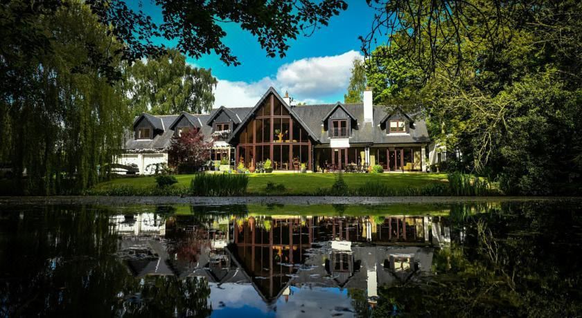 a large , two - story house with a gray roof and white walls is situated near a pond , surrounded by trees at Willowbeck Lodge Boutique Hotel