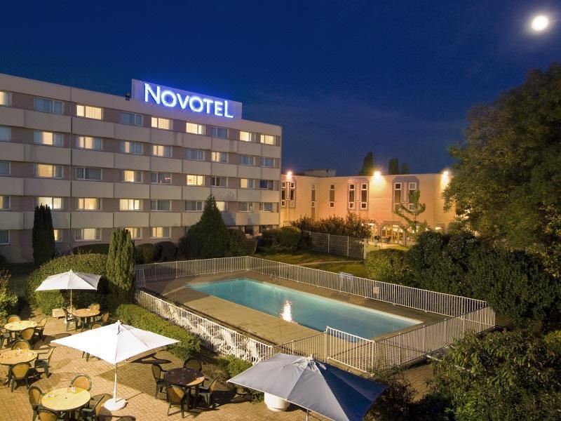 an outdoor pool area with a swimming pool , surrounded by chairs and umbrellas , and a hotel building in the background at Novotel Paris Nord Expo Aulnay