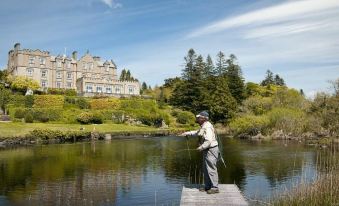 a man is standing on a dock , holding a fishing rod and casting a fishing line into a pond near a large house at Ballynahinch Castle Hotel